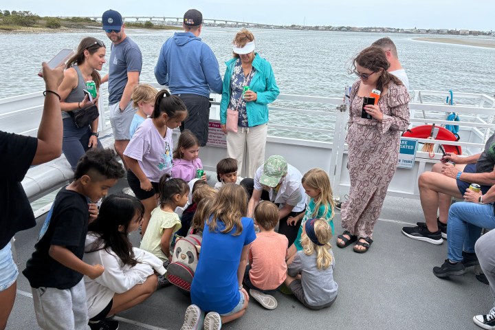 Children gathered around an adult on a boat, with adults standing nearby, water and bridge in the background.