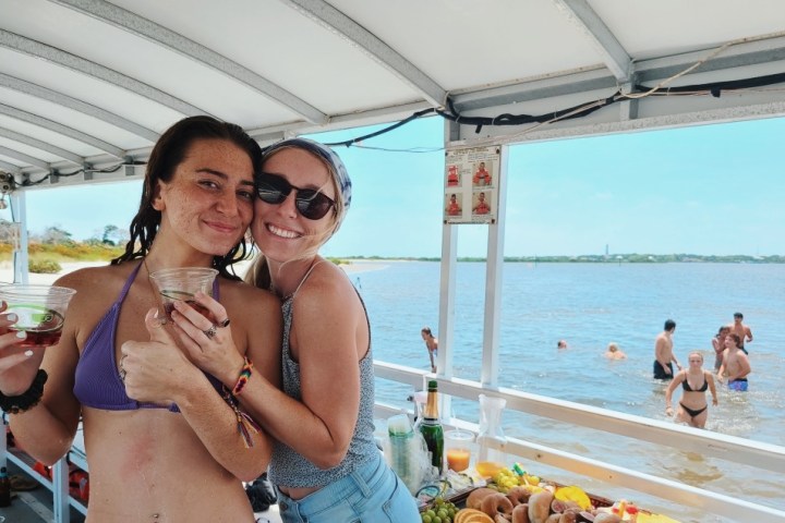 Two women on a boat, holding drinks, with people swimming in the background.