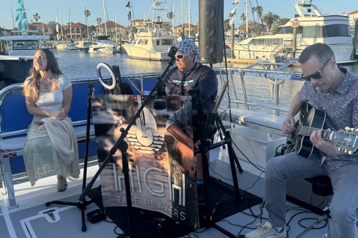 Musicians performing on a boat deck at sunset with docks and sailboats in the background.