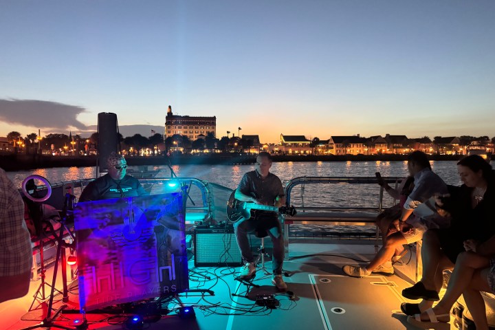 Musicians playing on a boat at sunset with city lights in the background.