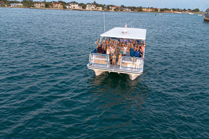 Group of people on a white boat in open water near a coastline with houses.