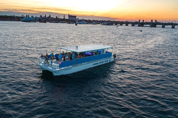 Boat with tourists on water at sunset, city skyline and bridge in background.