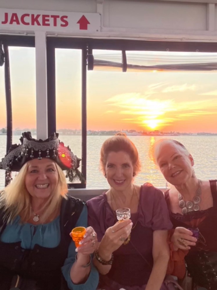 Three women in costumes holding small glasses on a boat with sunset view behind.