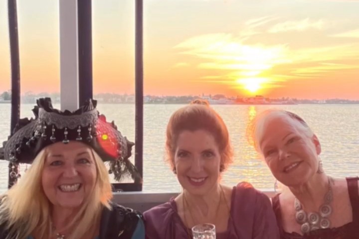 Three women in costumes holding small glasses on a boat with sunset view behind.