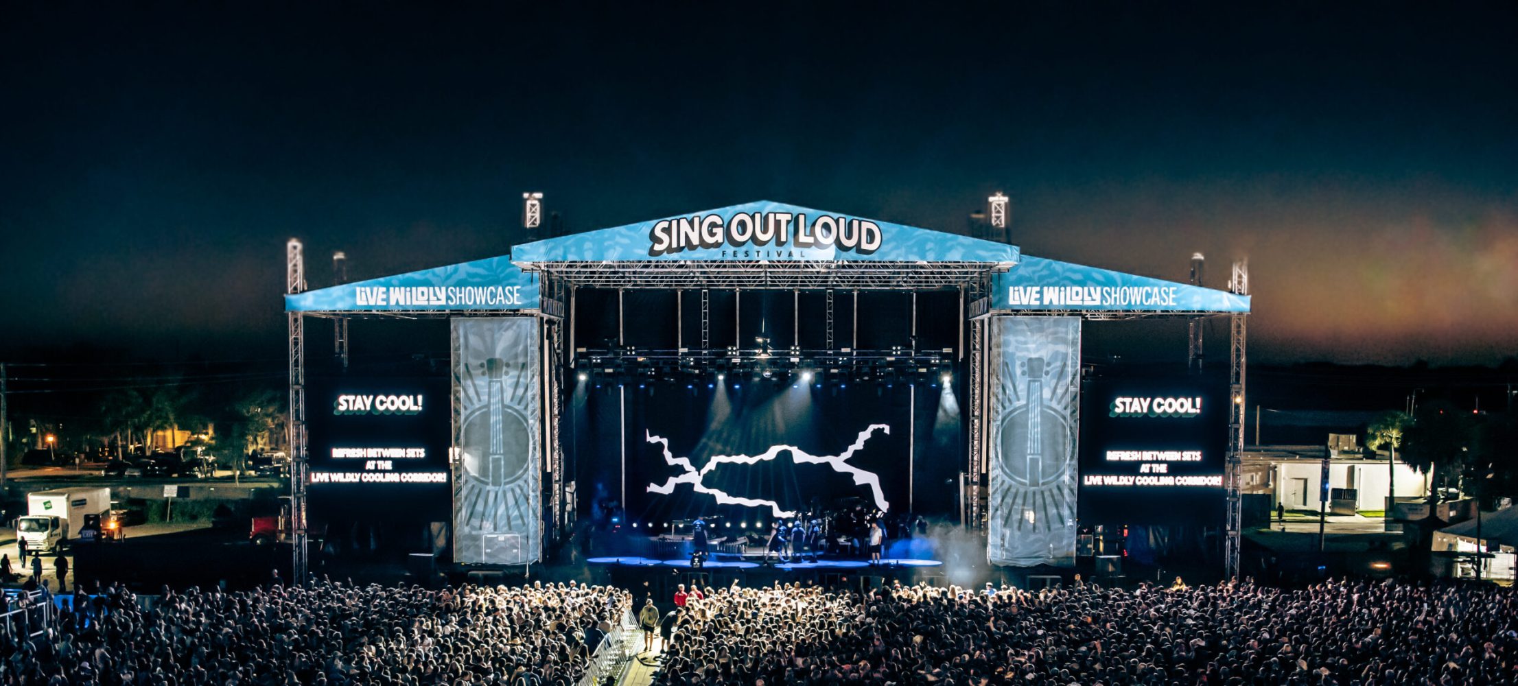 Crowd at outdoor night concert with stage lights and 'Sing Out Loud' banner.