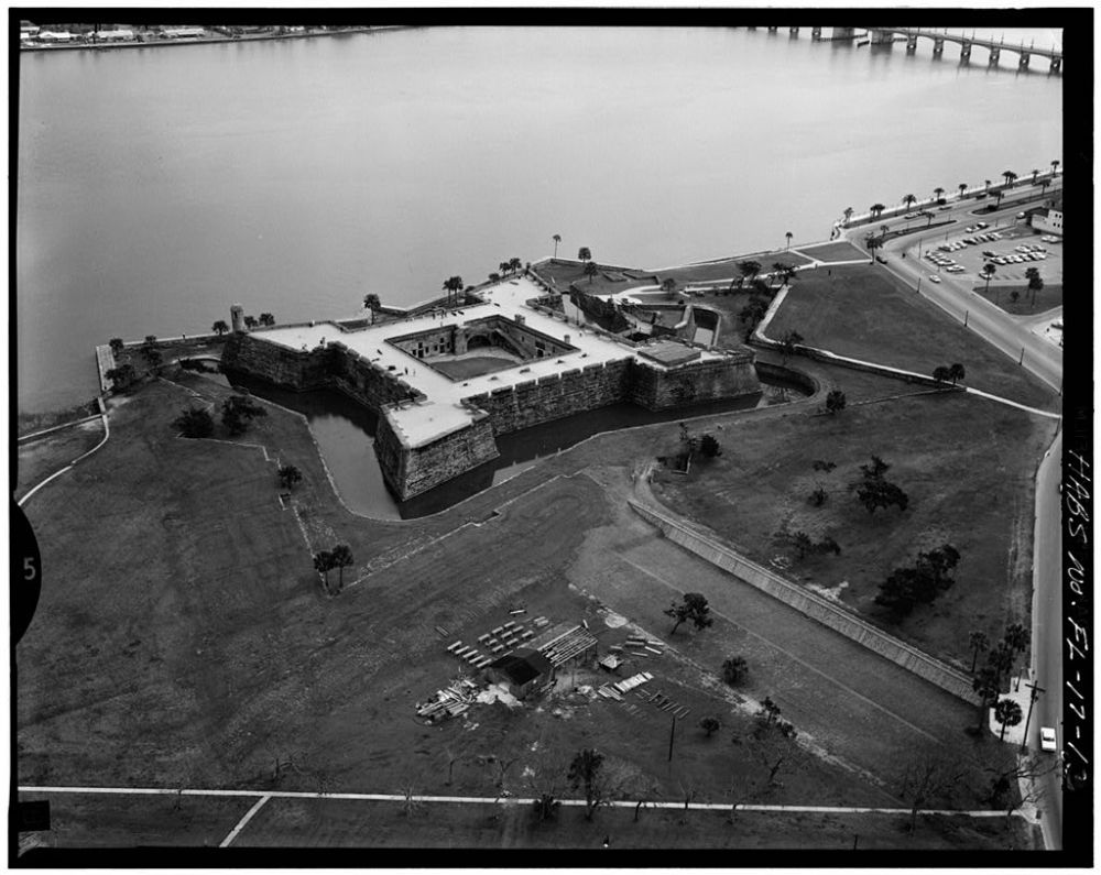Aerial view of a historic fort with a moat by a wide river, surrounded by open grassy areas and nearby road.