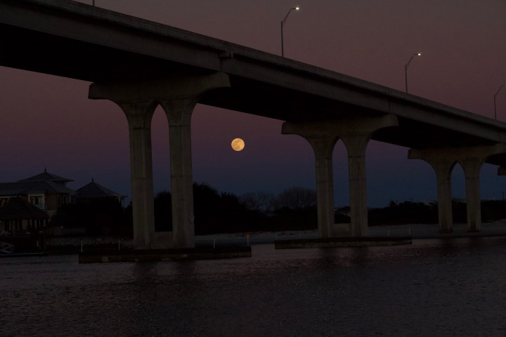 Full moon visible under a large bridge at dusk over a body of water.