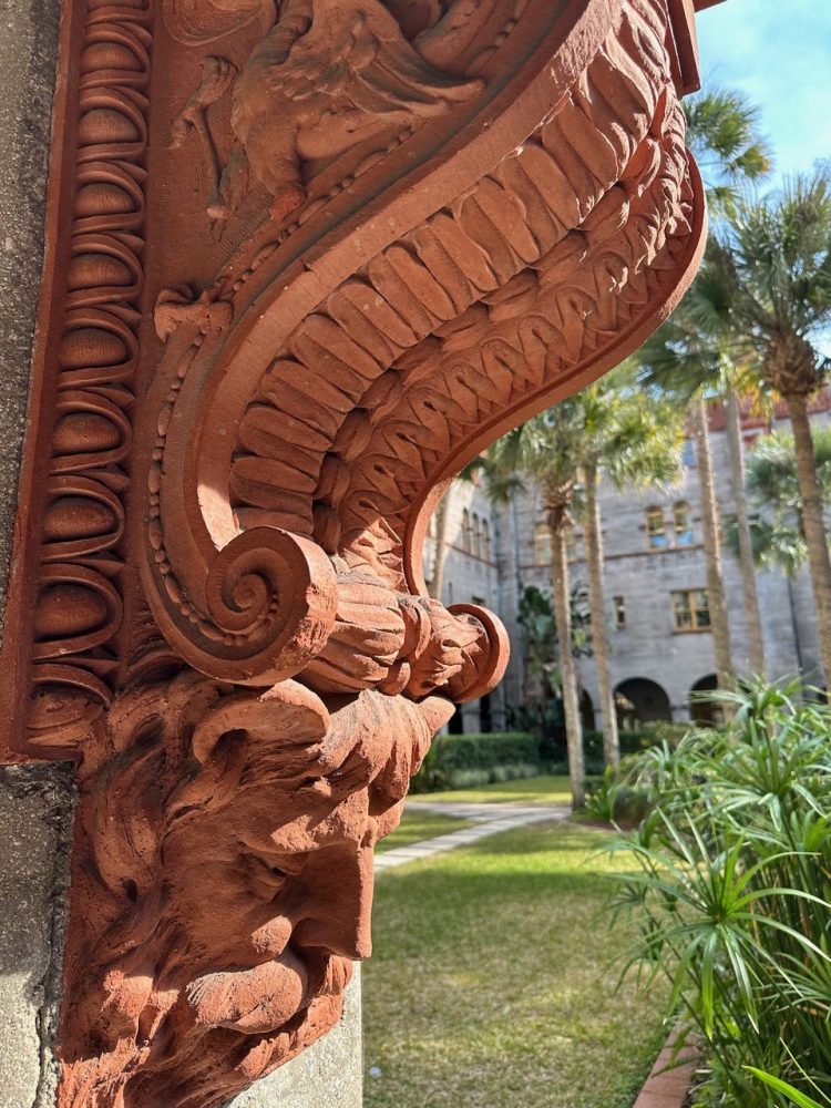 Ornate red stone column detail with a garden and building in the background.