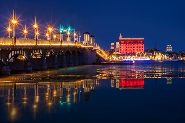 Illuminated bridge and buildings at night reflecting on water.
