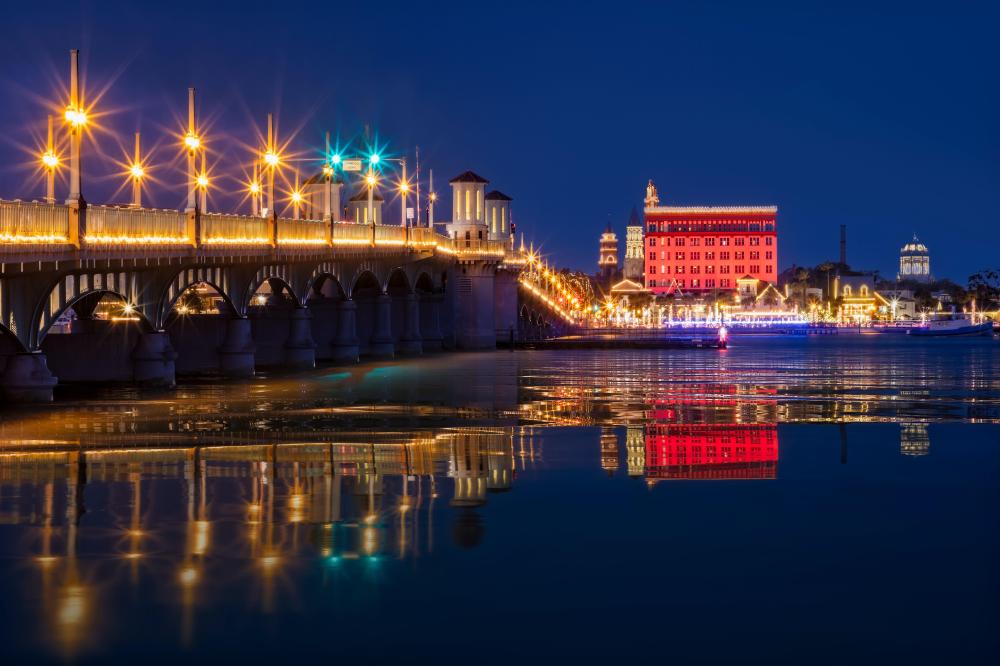 Illuminated bridge and buildings at night reflecting on water.