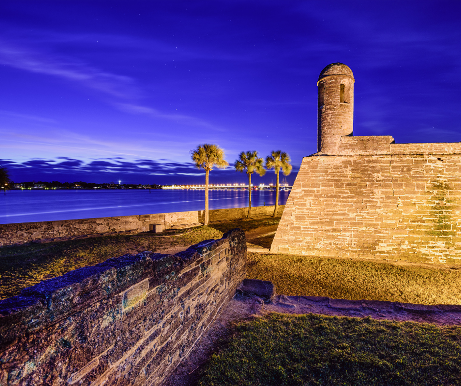 Historic stone fort by a river at dusk with palm trees and a lit bridge in the background.