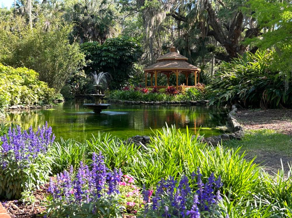Gazebo by pond with fountain, surrounded by lush greenery and purple flowers in foreground.