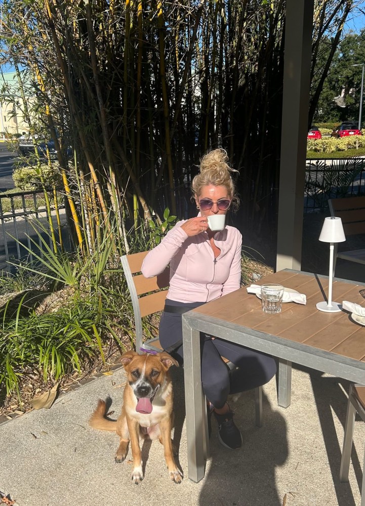 Person in sunglasses drinks coffee at an outdoor table with a dog sitting beside them.