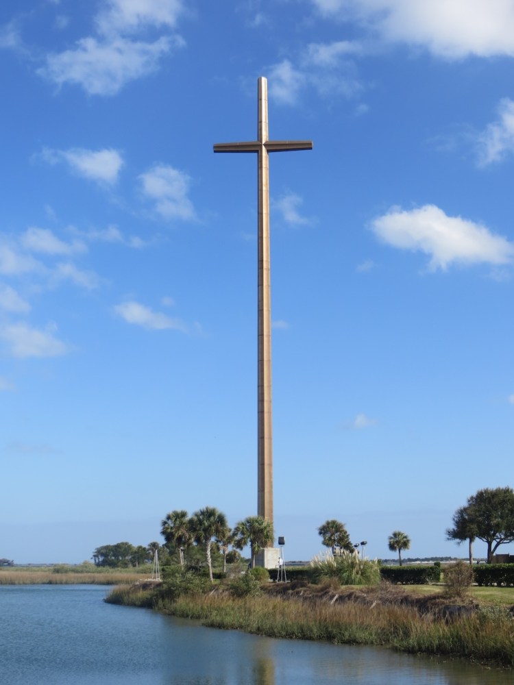 Tall cross near water with palm trees against a blue sky with clouds.