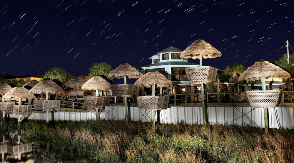 Night scene with tiki huts on stilts, starry sky with trails and a house in the background.