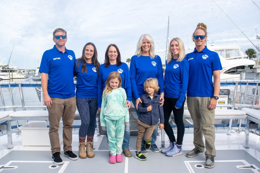 Group of eight people in blue shirts standing on a boat deck, with a marina in the background.