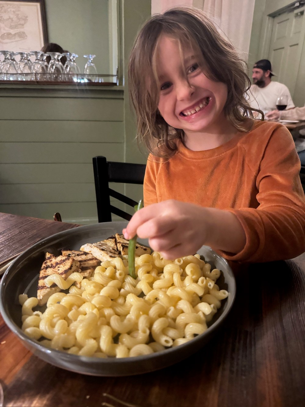 Child smiling at table with a plate of pasta and grilled chicken.
