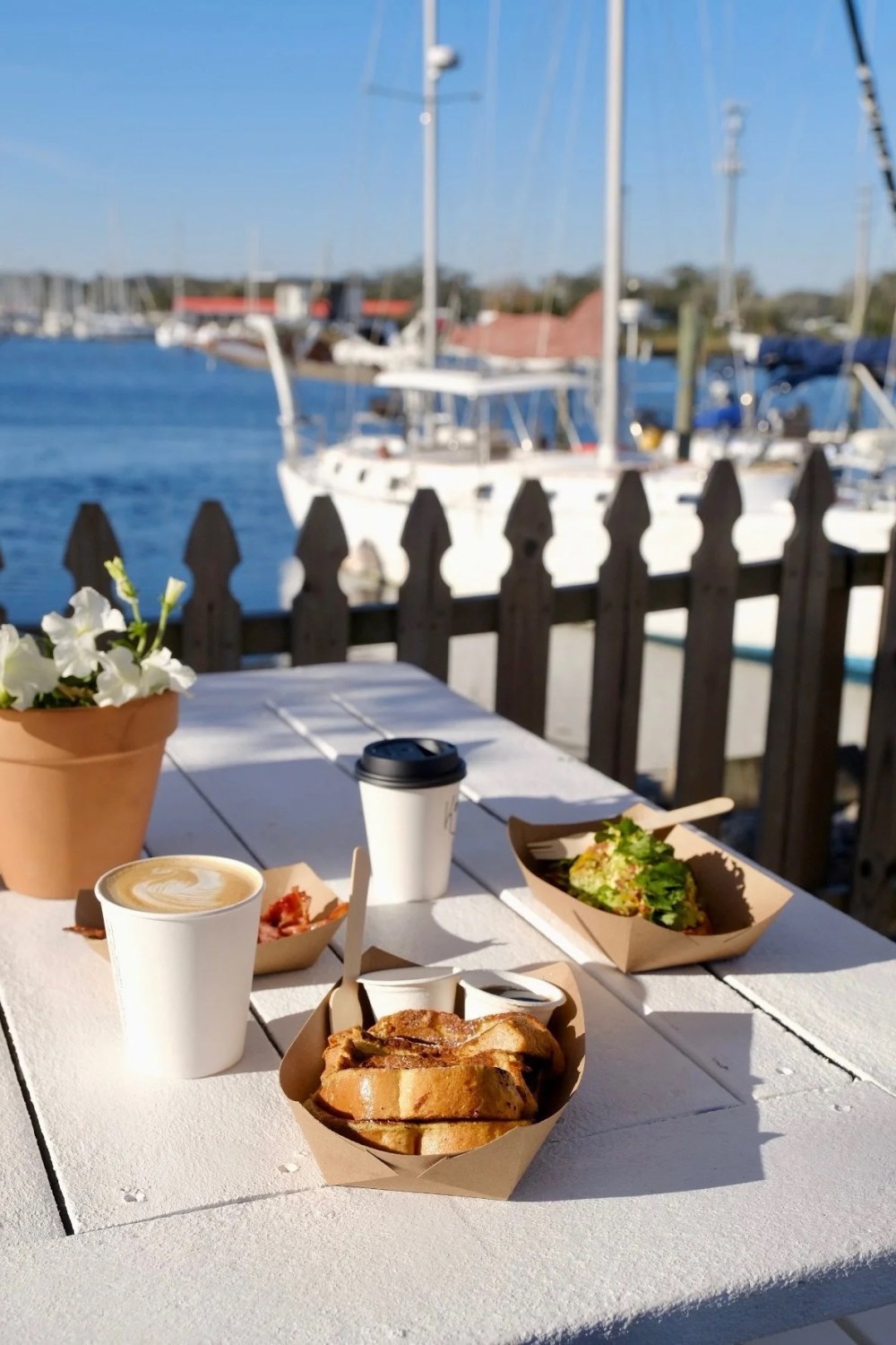 Outdoor dining table with pastries, coffee, and a potted plant by a marina.