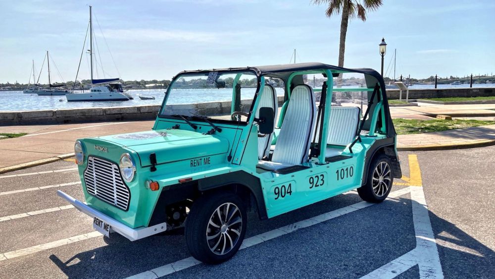 A turquoise open-top rental car parked by a waterfront with boats in the background.