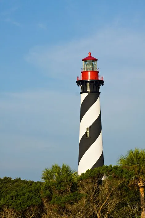 Black and white striped lighthouse with red top against blue sky and trees at base.