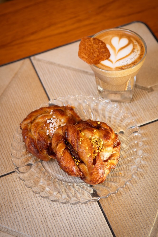 Two cinnamon buns on a glass plate with a latte art coffee in a glass beside them.