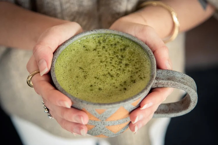 Hands holding a ceramic mug filled with frothy green matcha tea.