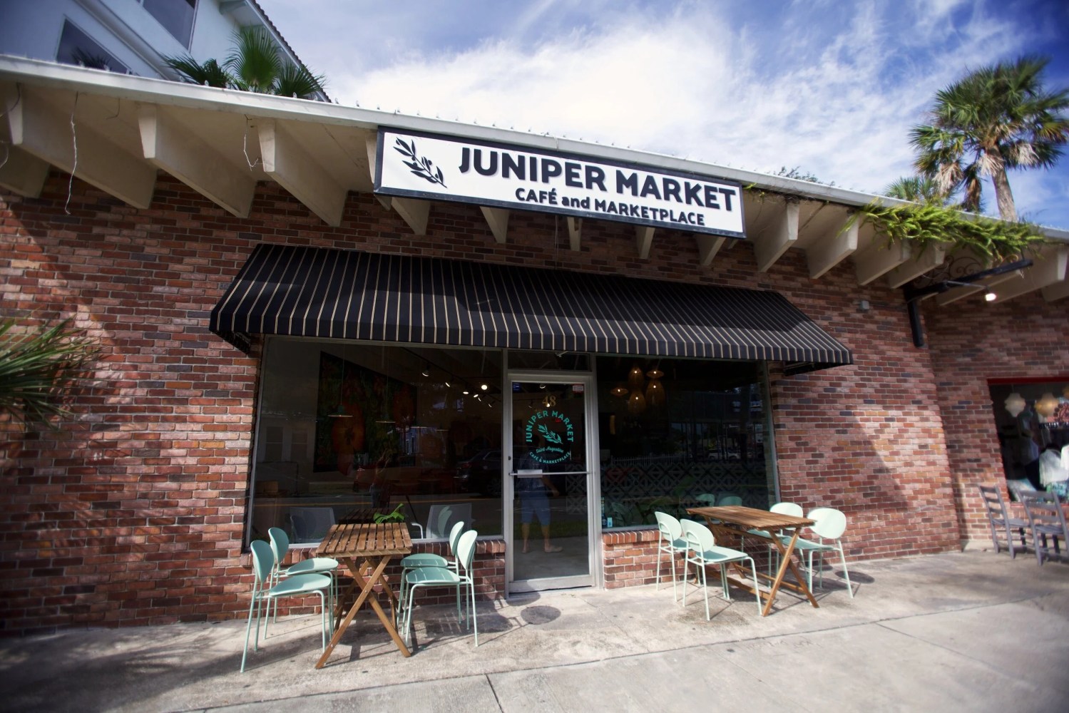 Brick cafe storefront with outdoor seating, striped awning, and sign reading 'Juniper Market'.