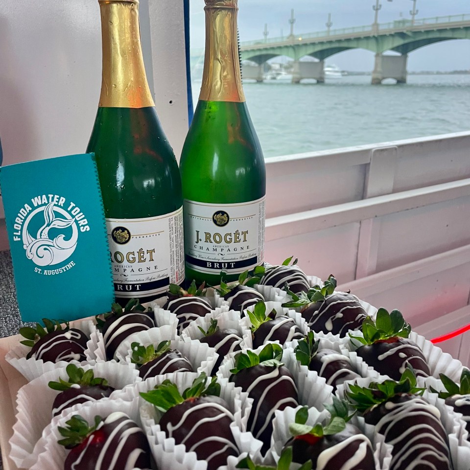 Two champagne bottles and chocolate strawberries on a boat with a bridge in the background.
