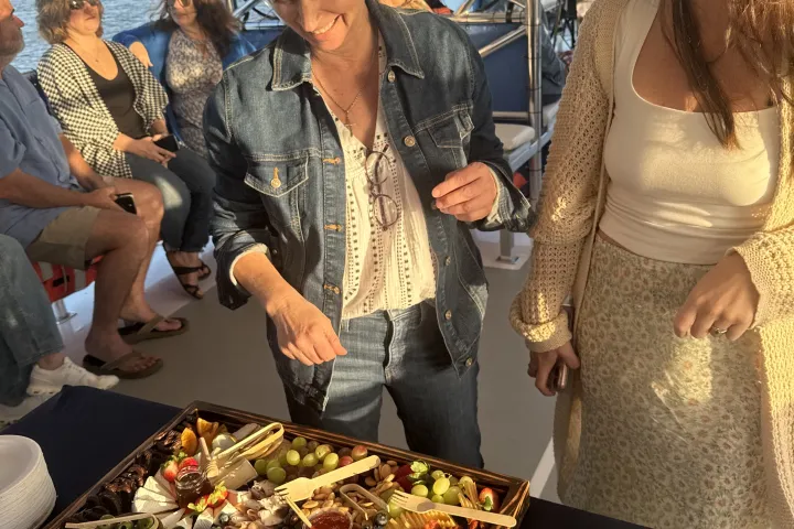 Two women smiling near a charcuterie board on a boat with seated passengers and water view.