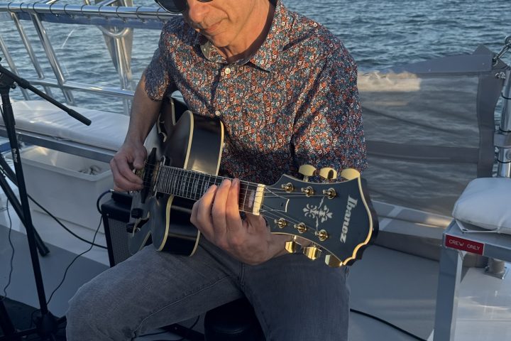 Man playing guitar on a boat with water in the background at sunset.