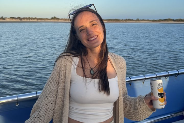 Smiling woman on a boat holding a drink, with water and shoreline in the background.