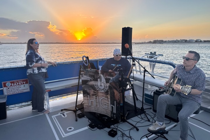Three musicians perform on a boat at sunset with water and land in the background.