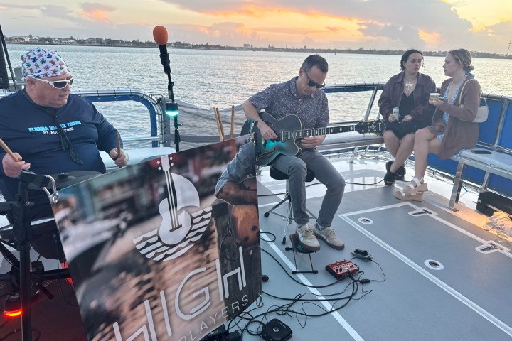 Musicians playing guitar and drums on a boat at sunset with two people sitting nearby.