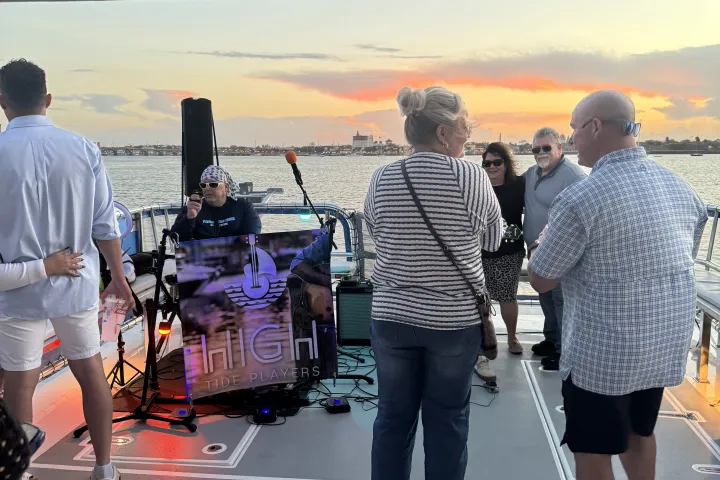 People on a boat listening to a musician at sunset with a 'High Tide Players' sign.