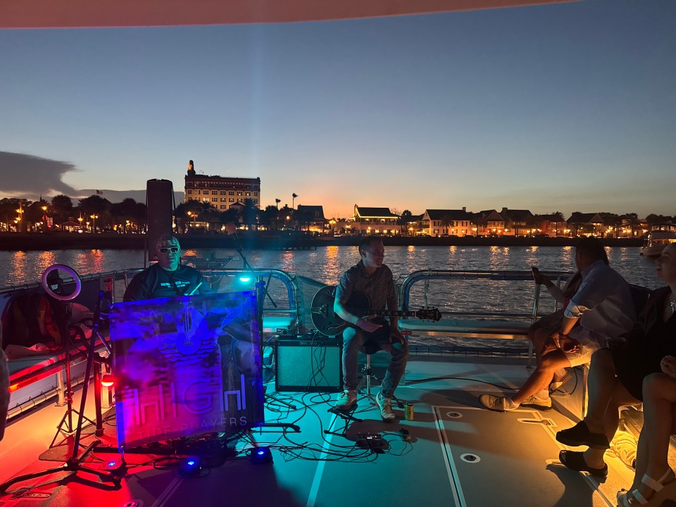 Evening boat music scene with city lights, musicians playing guitars, and seated guests.