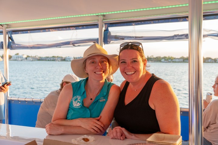 Two women smiling on a boat with water and houses in the background.