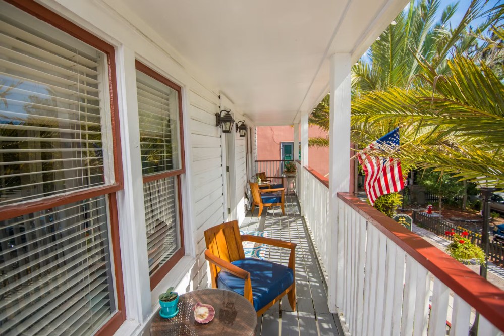 A wooden porch with chairs, table, and an American flag, surrounded by palm trees and white railings.