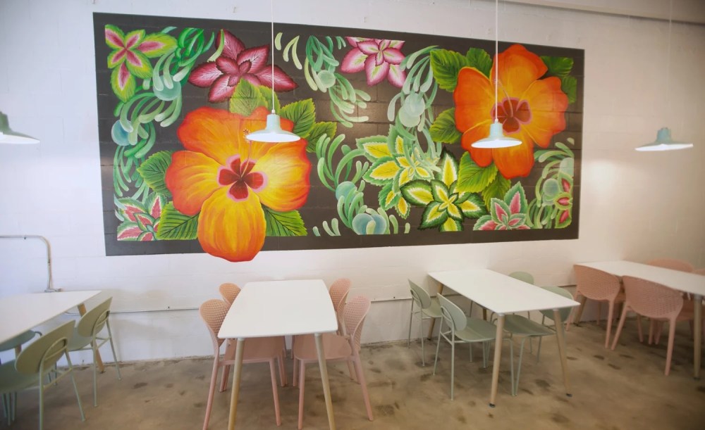 Cafe with colorful floral mural and white tables with pink and green chairs.