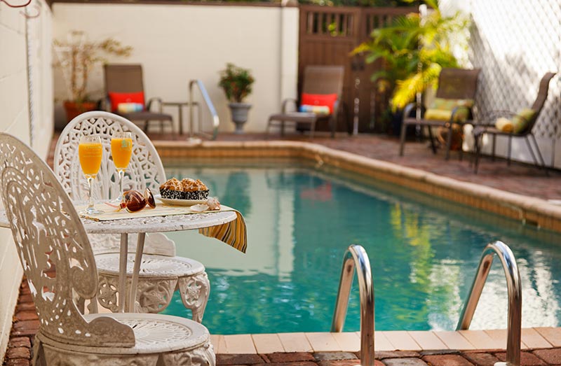 Poolside table with orange drinks, doughnuts, and sunglasses beside a swimming pool and lounge chairs.