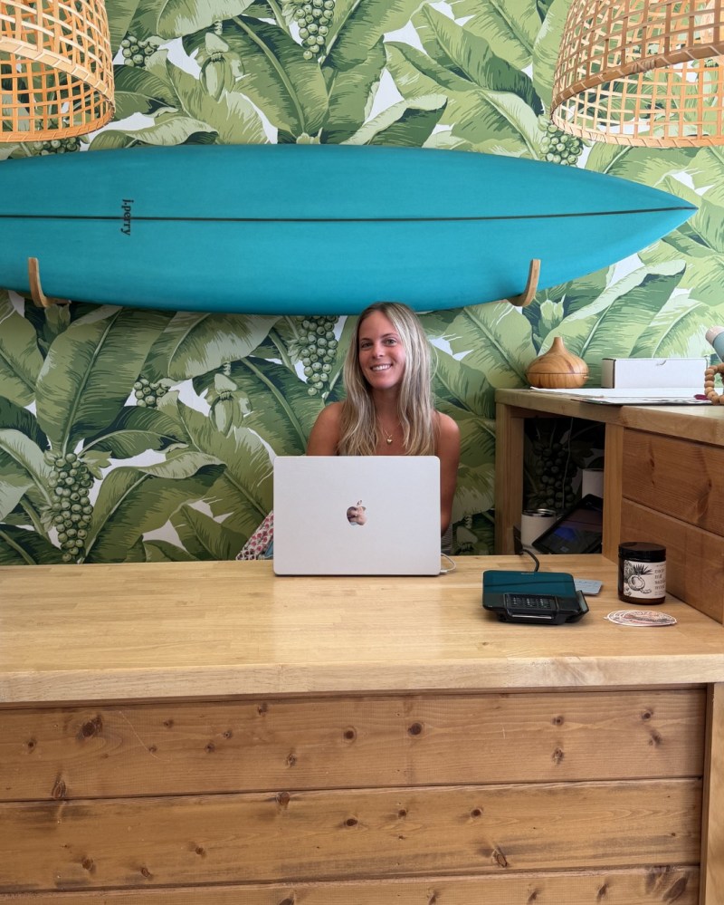 Woman at desk with laptop, surfboard on wall, tropical leaf wallpaper.