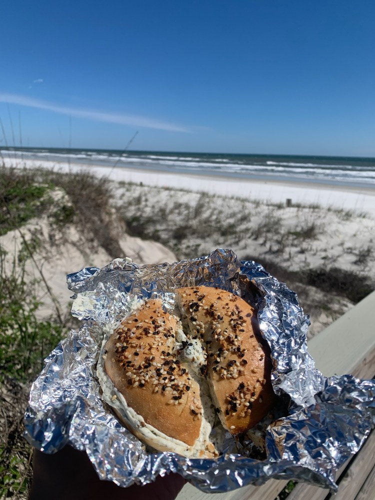 Bagel with cream cheese on foil, beach in background.