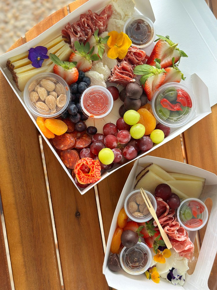 Two charcuterie boxes with fruits, nuts, cheese, and meats on a wooden table.