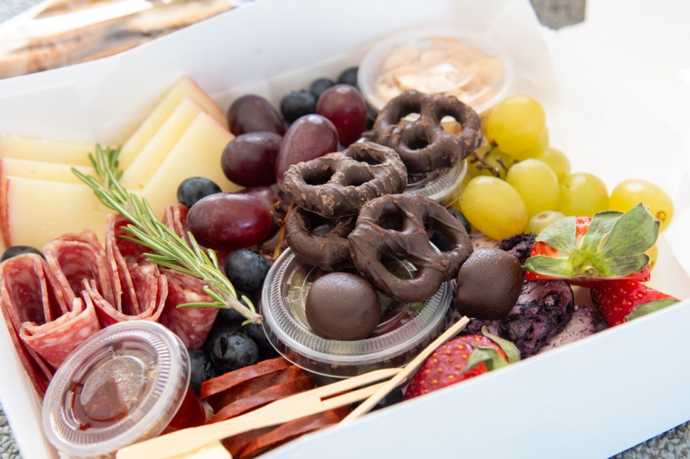 Box with fruit, cheese slices, salami, chocolate pretzels, and dips.