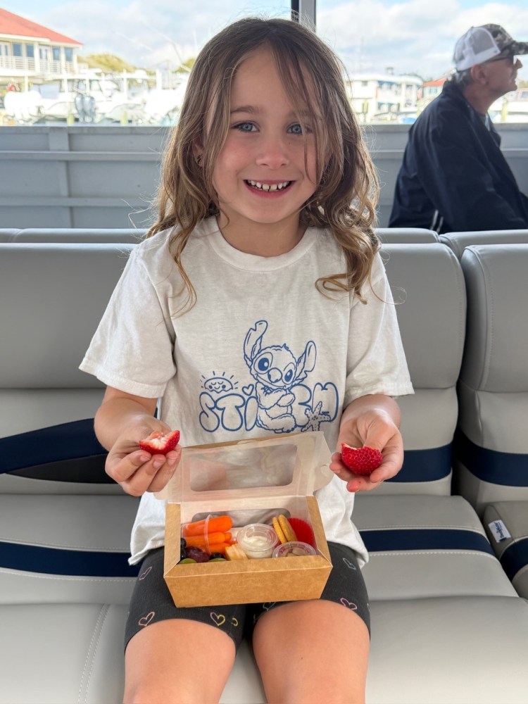 Smiling child holding strawberries, sitting with a snack box on a boat.