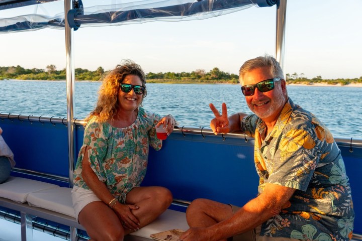 Two people smiling on a boat by the water, wearing sunglasses, with one person giving a peace sign.
