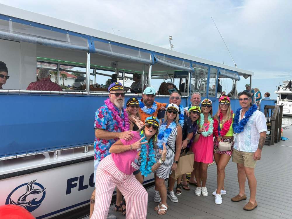 Group in colorful leis and captain hats posing on a dock by a blue boat.