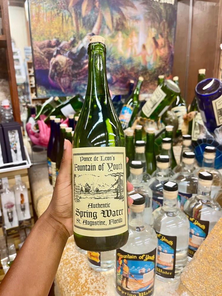 Hand holds a green bottle labeled 'Fountain of Youth Spring Water' among other bottles.