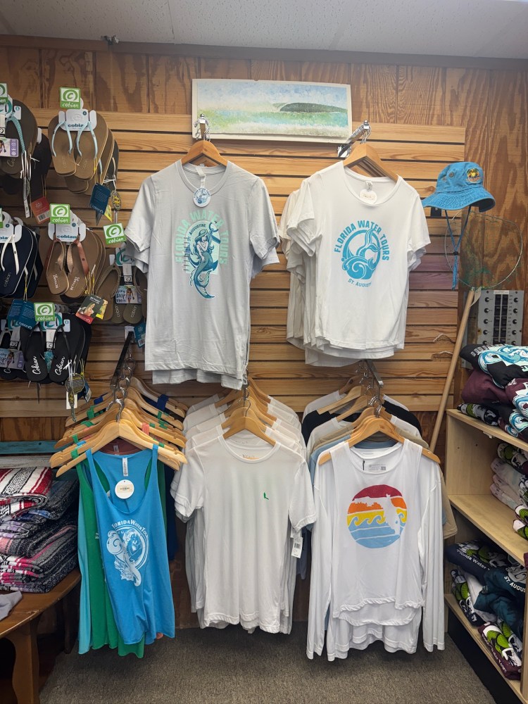 Wall display of t-shirts with beach designs, sandals, and a blue hat at a store.
