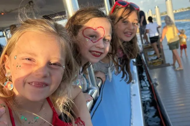 Girls leaning over boat railing, smiling on sunny day.