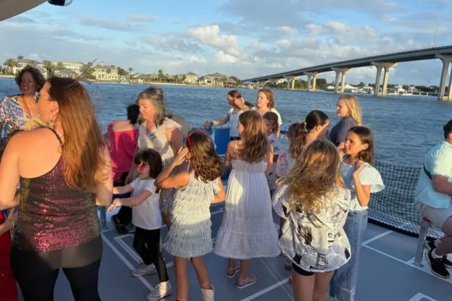 Group of people, including children, dancing on a boat with a bridge and water in the background.